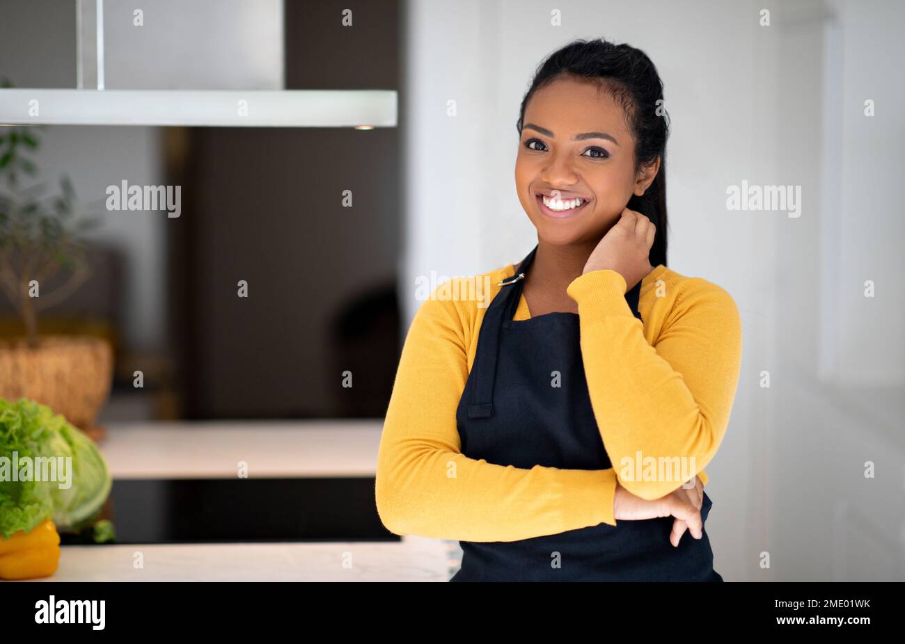 Smiling young african american lady cook in apron near table with vegetables in minimalist kitchen interior Stock Photo