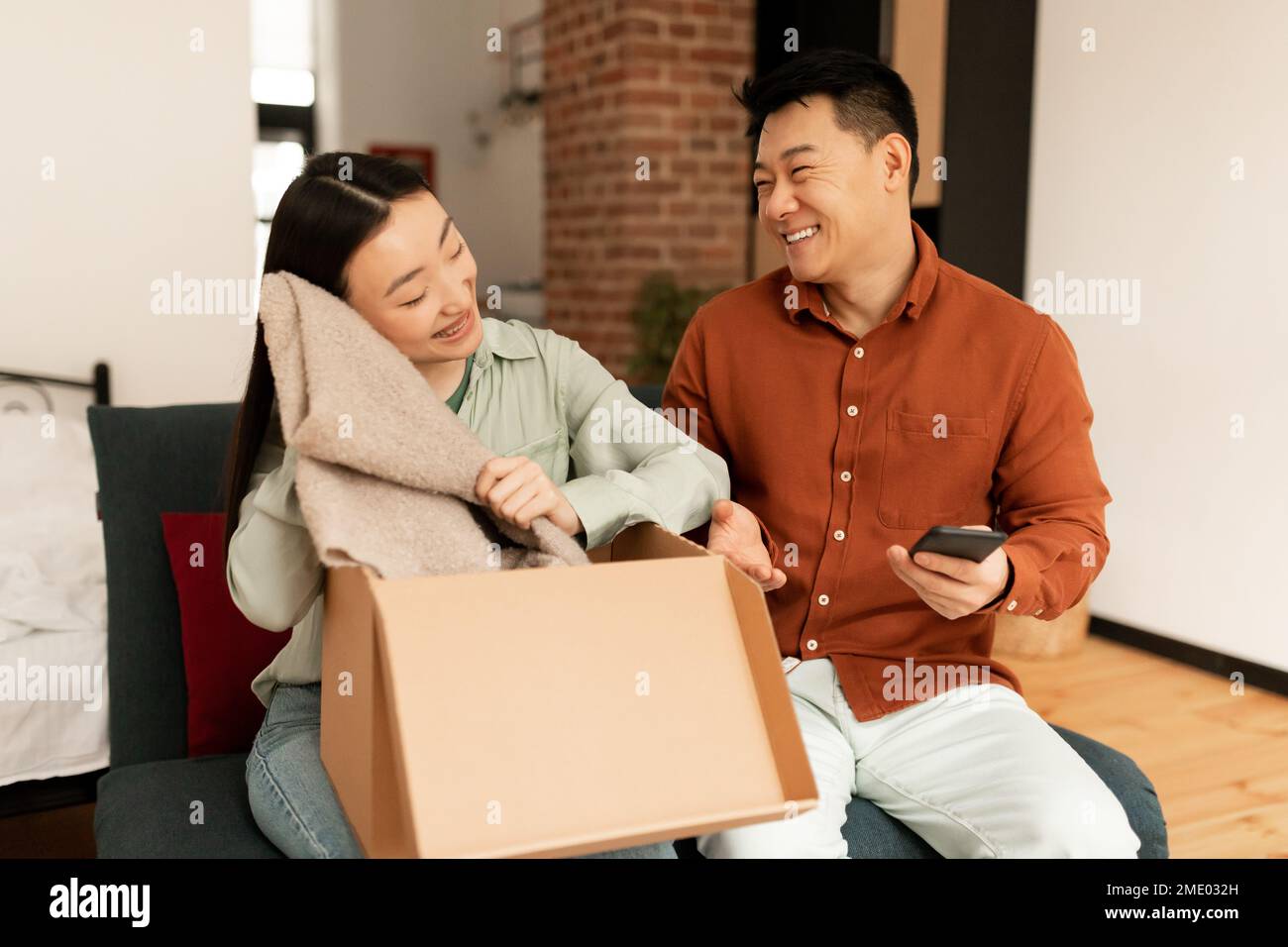 Successful online shopping. Happy asian female customer unpacking delivered box from store, sitting with husbnad on sofa Stock Photo