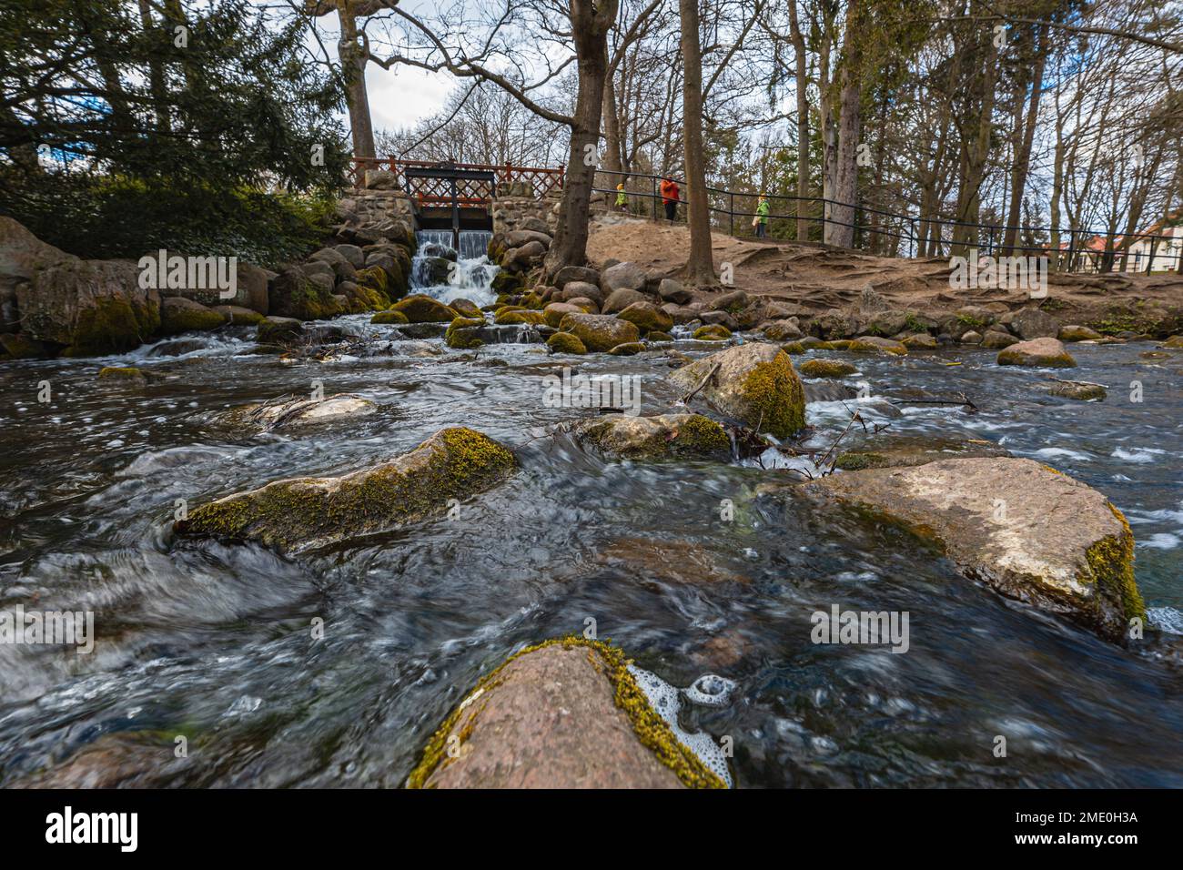 Small waterfall next to path in small Oliwski park with small wooden bridge over it and small pond full of big stones Stock Photo