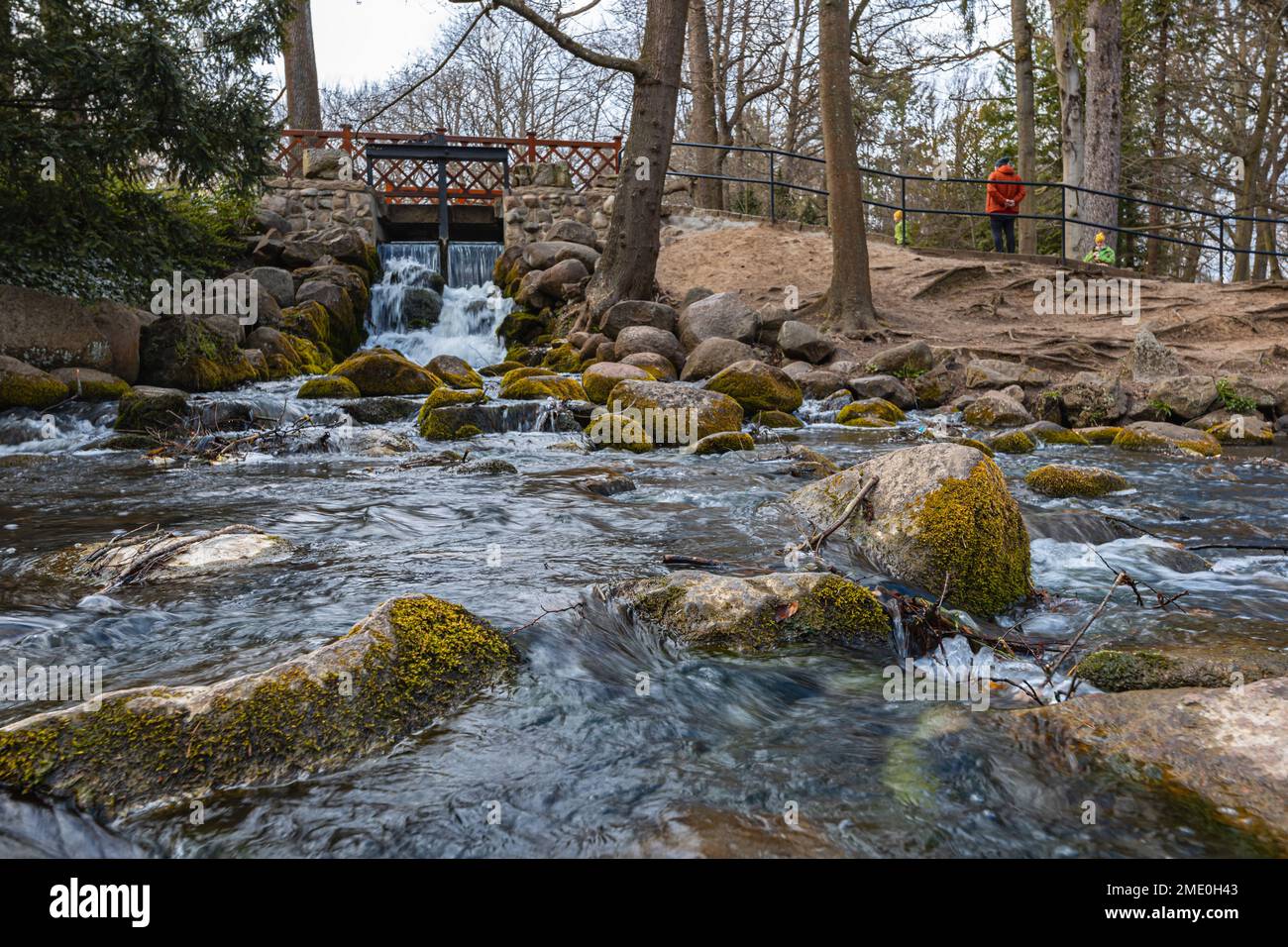 Small waterfall next to path in small Oliwski park with small wooden bridge over it and small pond full of big stones Stock Photo