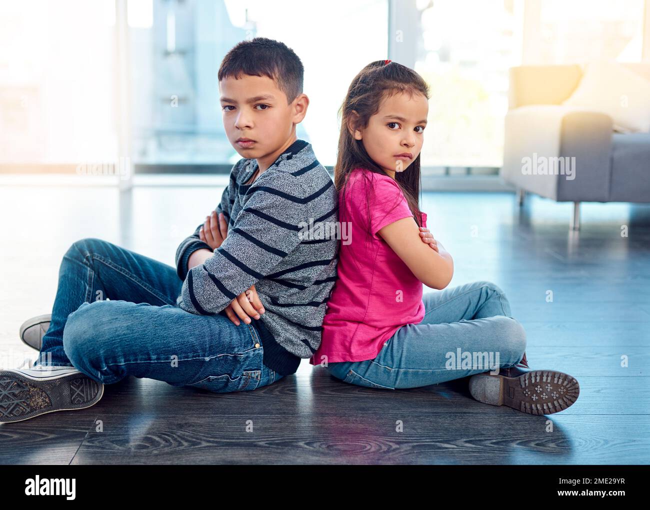 Good luck trying to cheer them up. Portrait of two naughty young children sitting down on the floor with their backs facing each other at home. Stock Photo