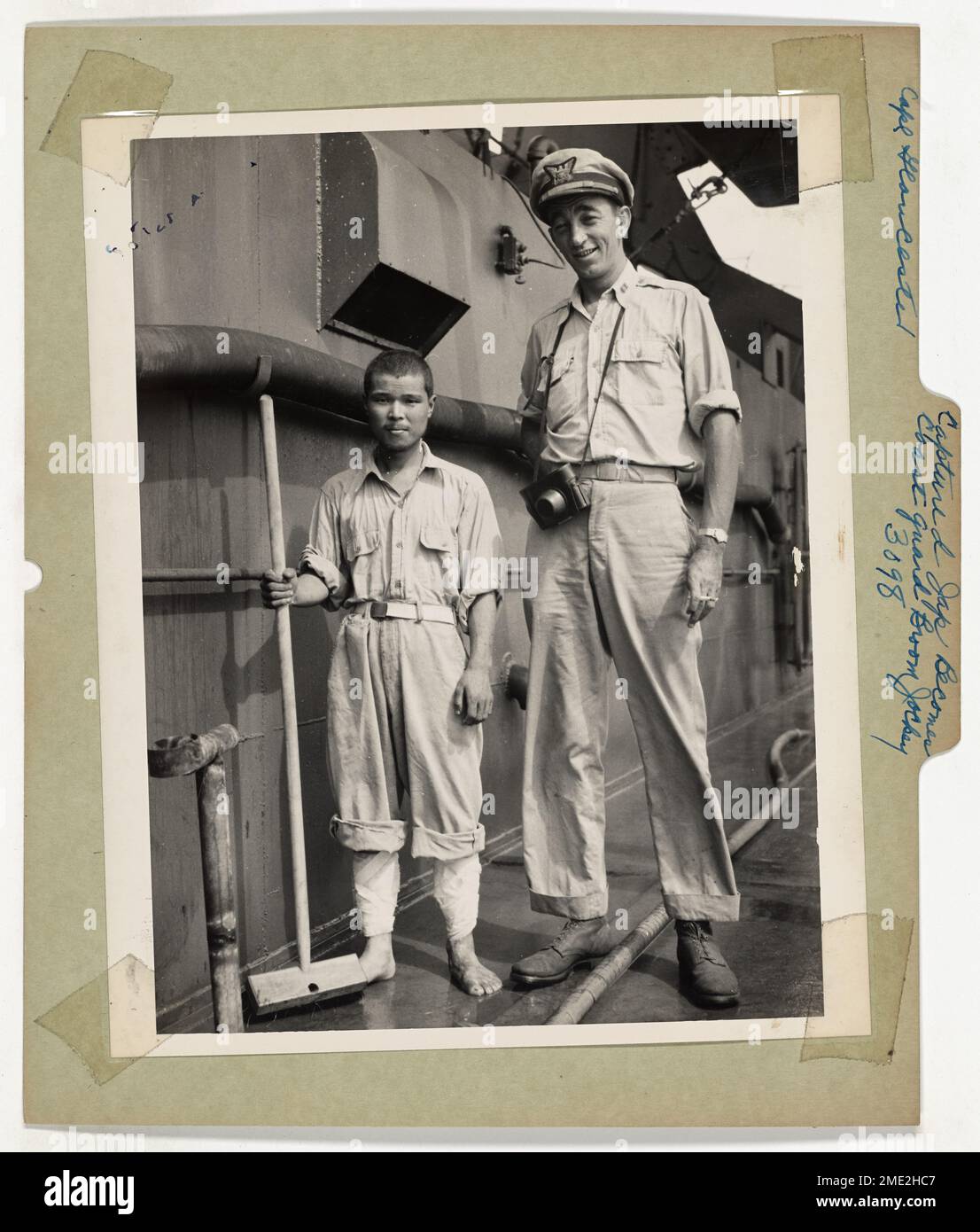 Photograph of Coast Guardsman with Japanese Prisoner of War. Captured Jap Becomes Coast Guard Broom Jockey. Coast Guard Lieut. Robert Edge towers above a captured Jap, who has been assigned to pushing a broom aboard a Coast Guard-manned invasion transport. The Nip is one of those who preferred life to 'immortal death' in the service of his Emperor, and became a prisoner of war in the Cape Gloucester, New Britain, invasion. Lieut. Edge's home is at 235 East 22nd Street, New York City. Stock Photo