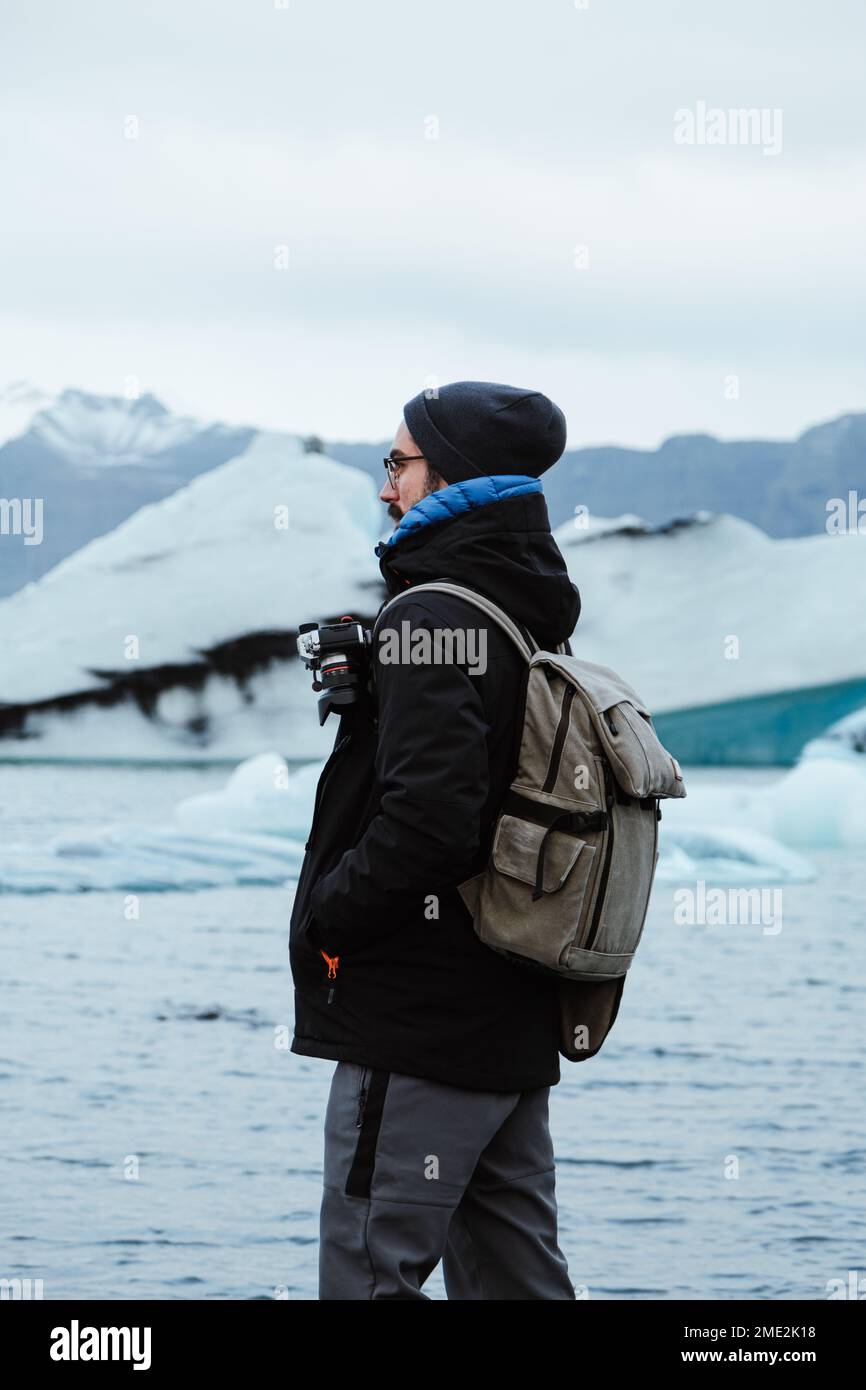 Side view of male tourist photographer in outerwear with camera and backpack holding hands in pockets while standing on stony coast covered with ice a Stock Photo
