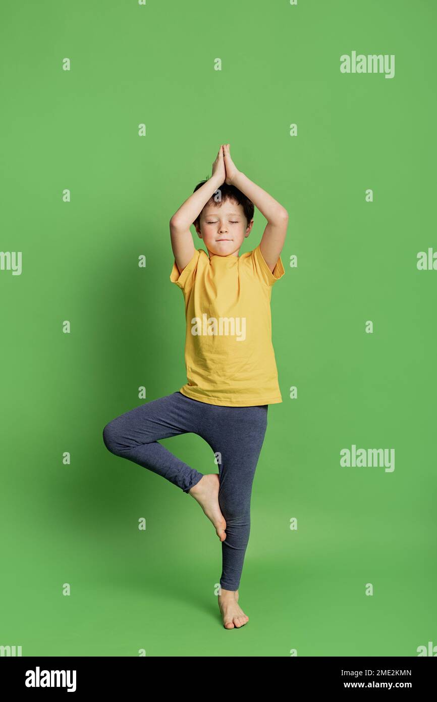 Full body of barefoot child in sportswear standing in Vrikshasana while practicing yoga in studio on green background Stock Photo
