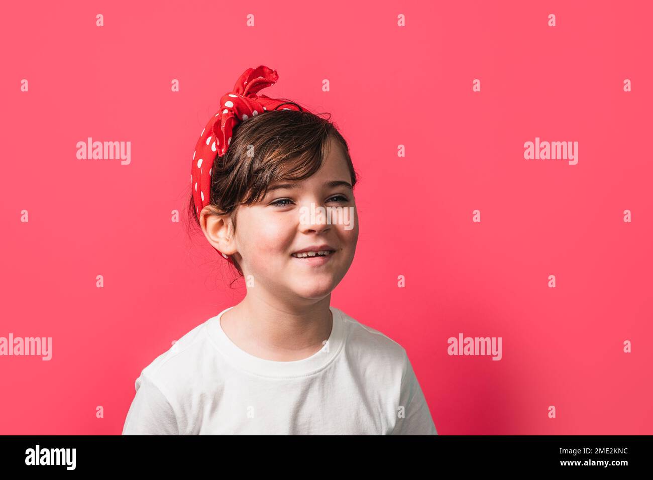 Adorable little girl in white t shirt with red polka dot headband looking away while standing against pink background Stock Photo