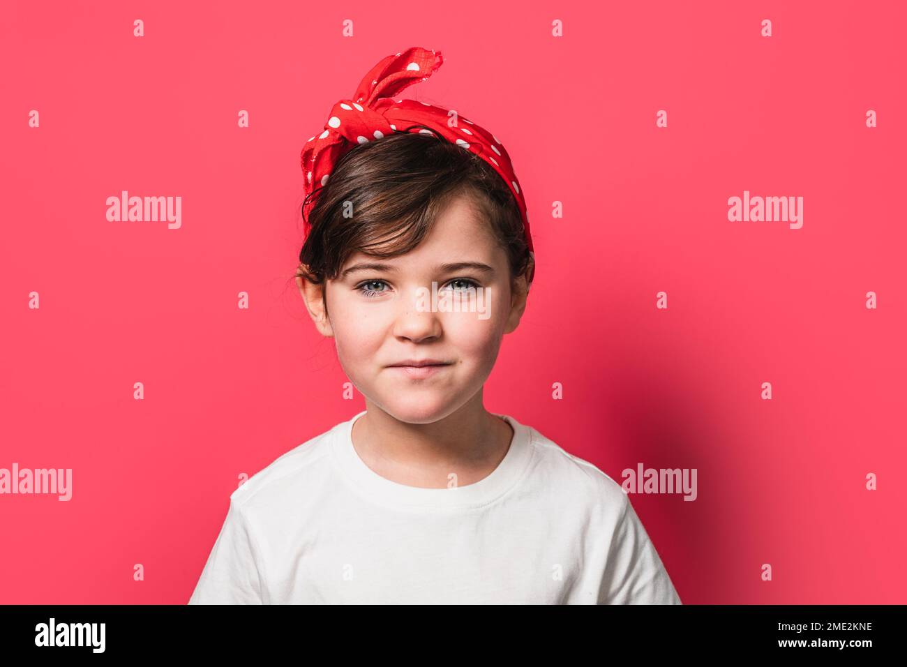 Adorable little girl in white t shirt with red polka dot headband looking at camera while standing against pink background Stock Photo