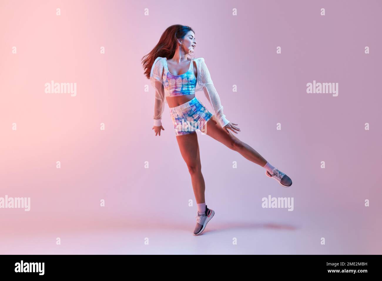 Young slim female in tight sportswear raising leg while dancing against pink background in studio Stock Photo