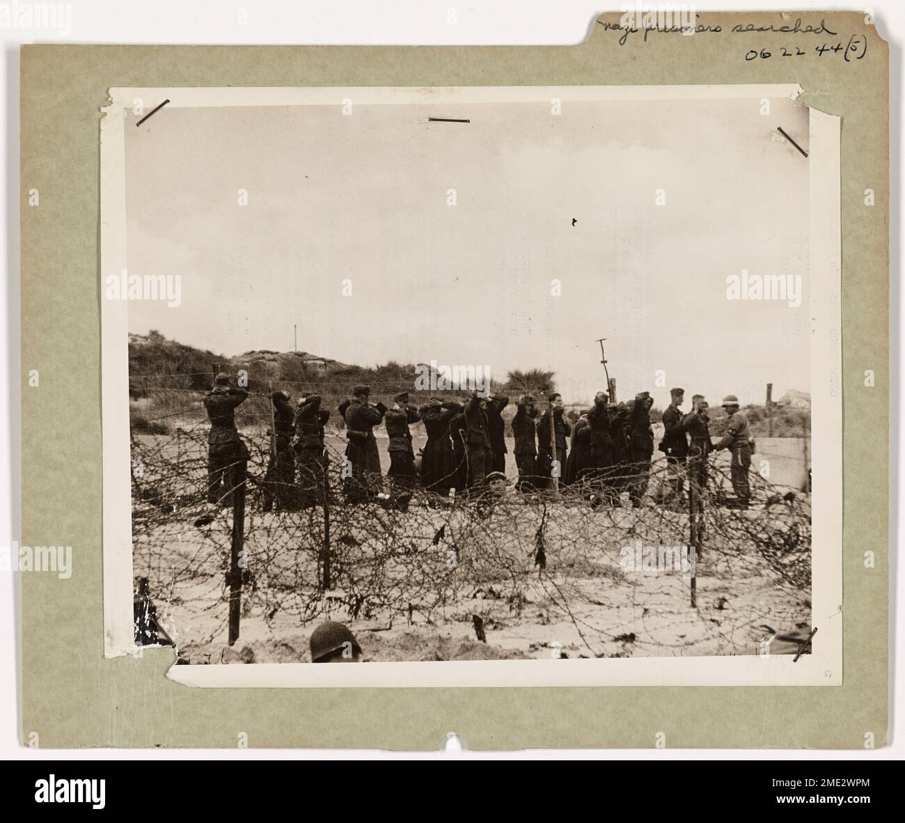 Nazi Prisoners Searched on French Beach. Their hands grasping the backs of their necks, a group of Nazi prisoners line up back of the barbed wire to be searched by an M.P. before they are transferred to landing craft and taken to England. This picture was made by a Coast Guard Combat Photographer who went in with the invaders. Stock Photo