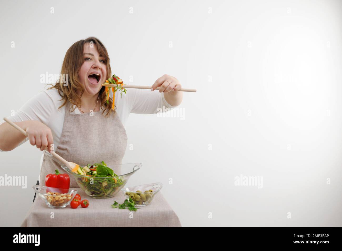 funny cheerful woman eating salad on white background she opens mouth wide bulges eyes large wooden spoon stuffs portion of food on table ingredients space for text weight loss ad Stock Photo