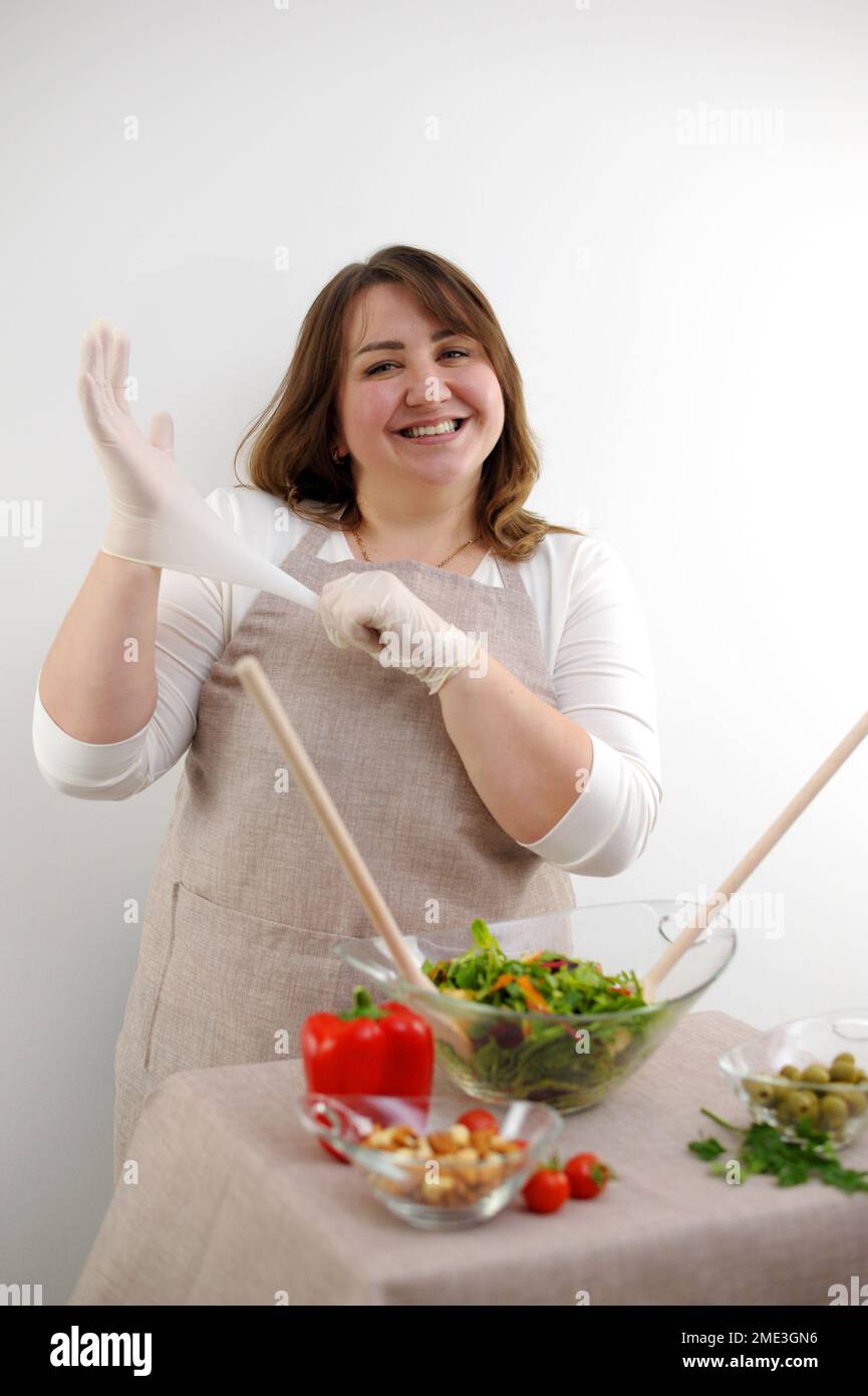 beautiful plump woman She cooks food in gloves wearing gloves looks into frame smiles concept of cleanliness neatness healthy attitude to cooking restaurant food on white background beige apron Stock Photo