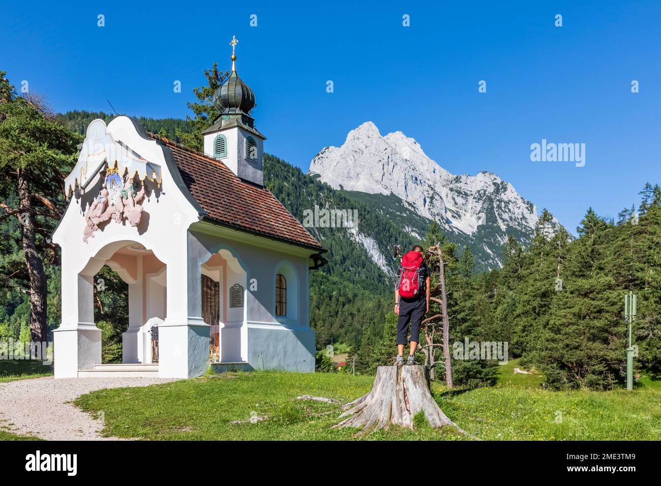 Germany, Bavaria, Female hiker standing on top of tree stump in front of Kapelle Maria Konigin Stock Photo