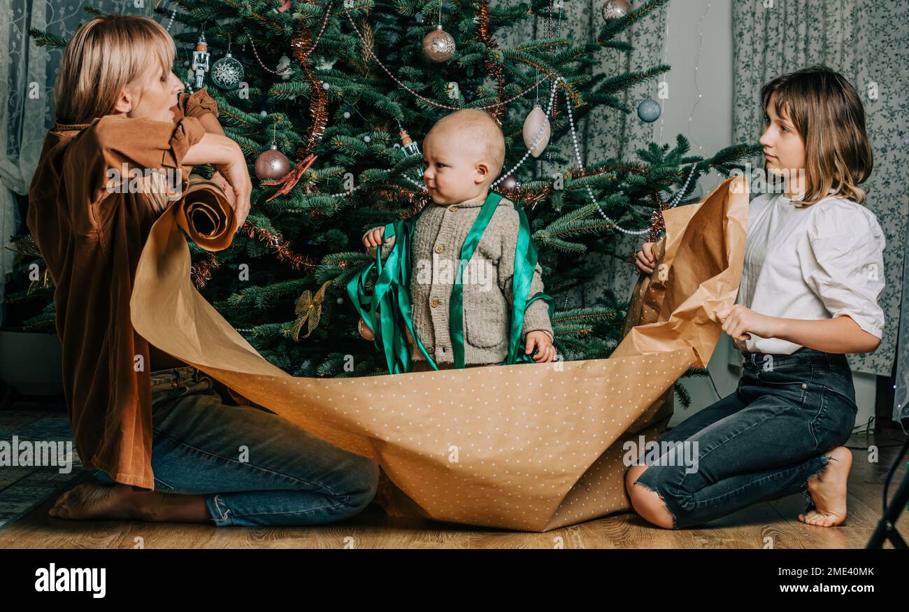 Mother and daughter holding wrapping paper with baby boy standing in front of Christmas tree Stock Photo