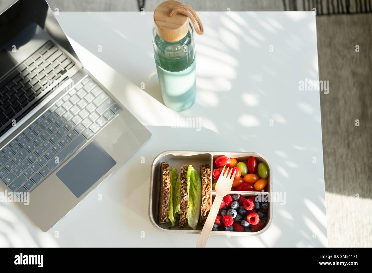 Rye bread, berries and salad with disposable fork in lunch box on table by water bottle and laptop Stock Photo