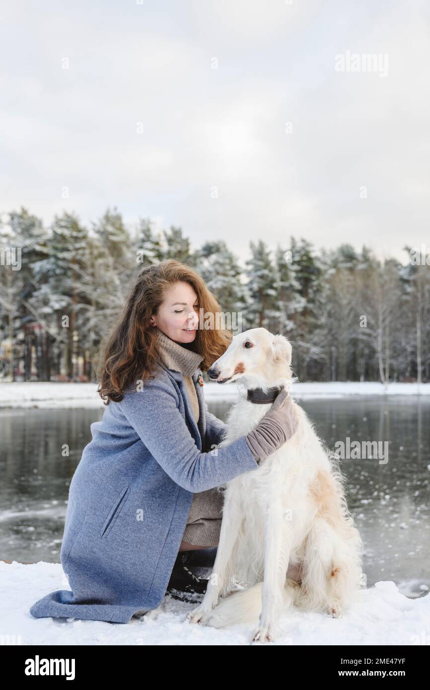 Mature woman stroking greyhound dog in front of frozen lake Stock Photo