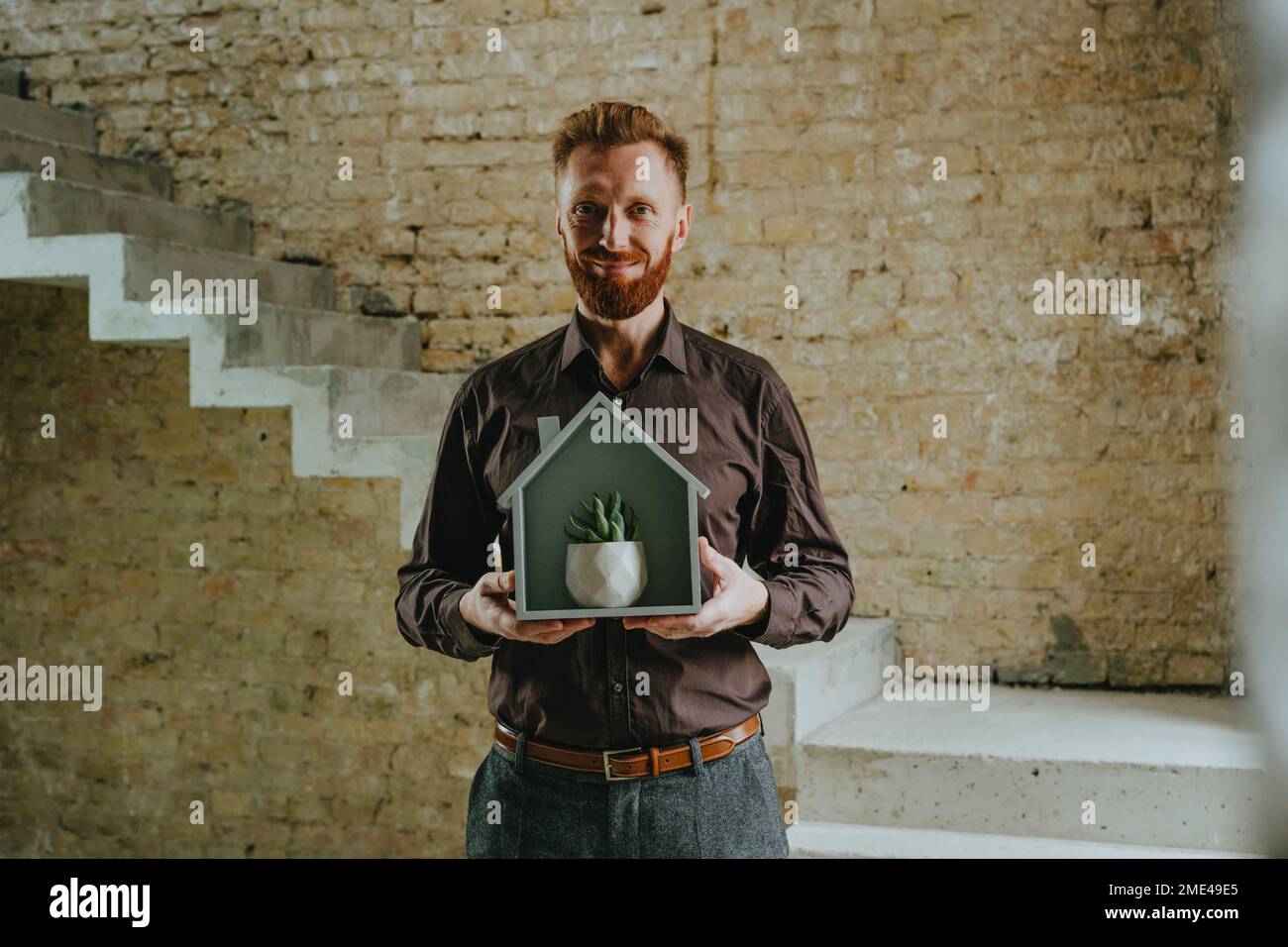 Happy businessman holding house model with plant in front of brick wall ...