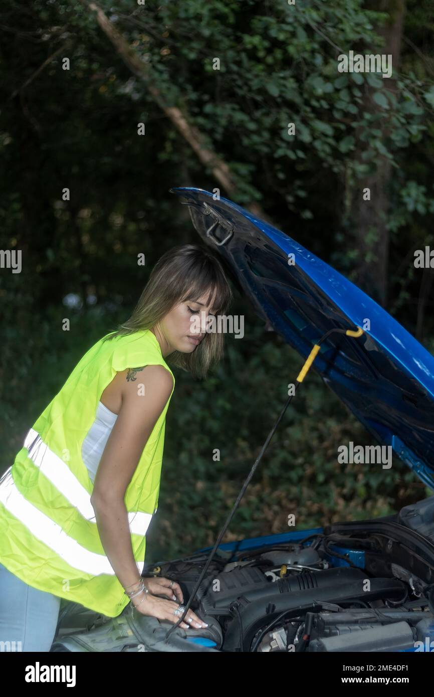 beuty woman in reflective vest looking at engine of broken down car Stock Photo
