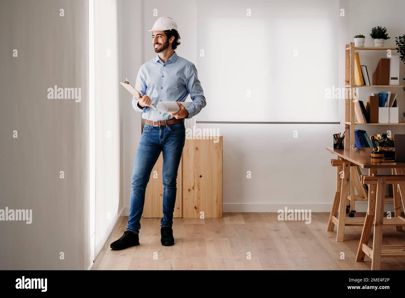 Contemplative smiling architect standing with blueprints in office Stock Photo