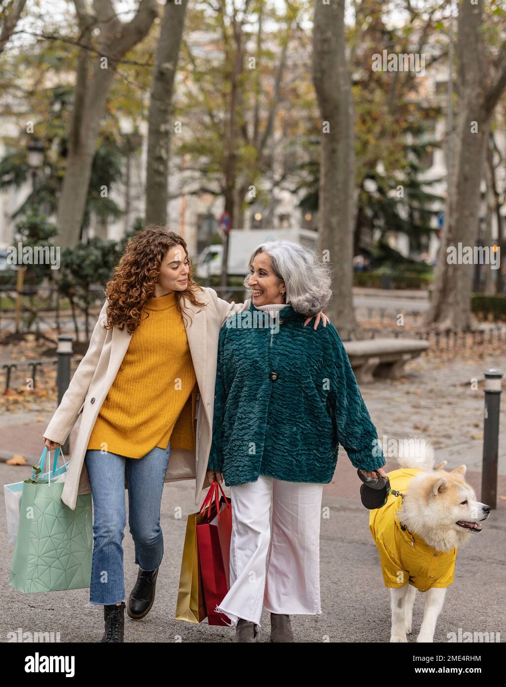 Happy granddaughter and grandmother holding shopping bags walking with dog at footpath Stock Photo