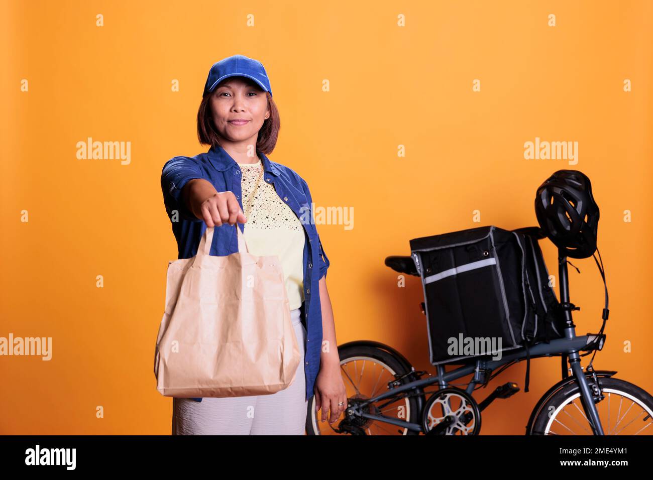 Pizzeria delivery worker wearing blue uniform while delivering fast food order to customer during lunch time. Restaurant employee standing in studio with yellow background. Takeout service concept Stock Photo