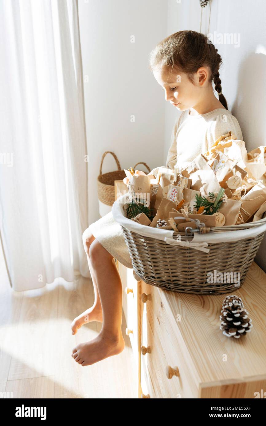 Girl opening gifts sitting on cabinet by basket of advent calendar at home Stock Photo