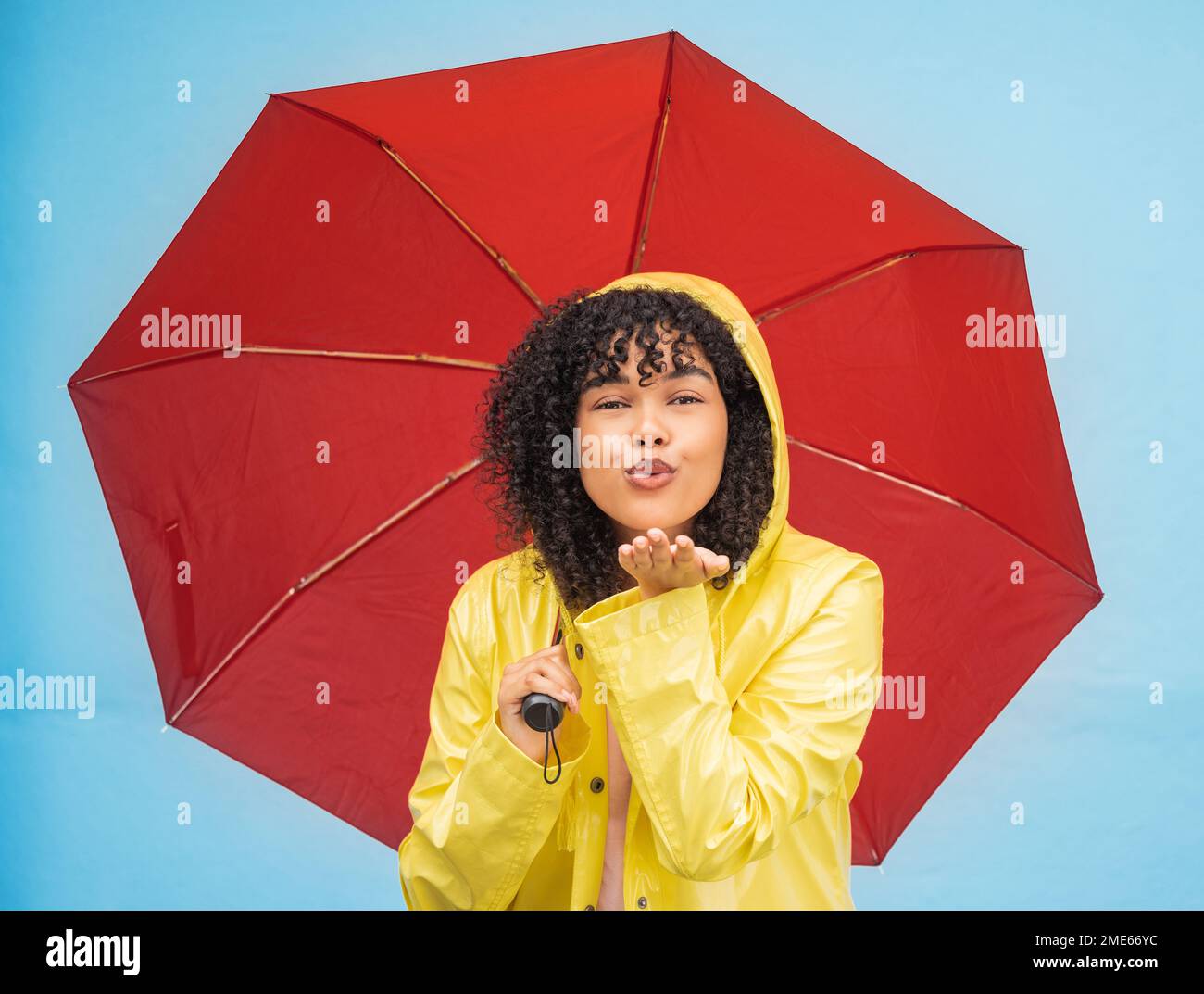 Woman, portrait or blowing kiss and rain umbrella on isolated blue background in Brazil city love or support. Black person, raincoat or flirty student Stock Photo