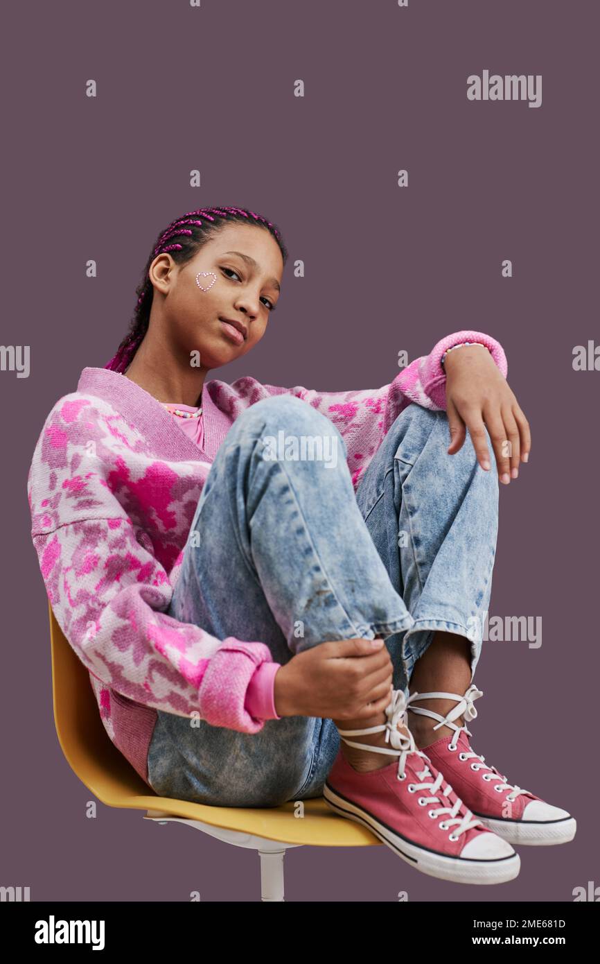 Vertical full length portrait of black teenage girl wearing trendy casual clothes and posing sitting on chair with feet up Stock Photo