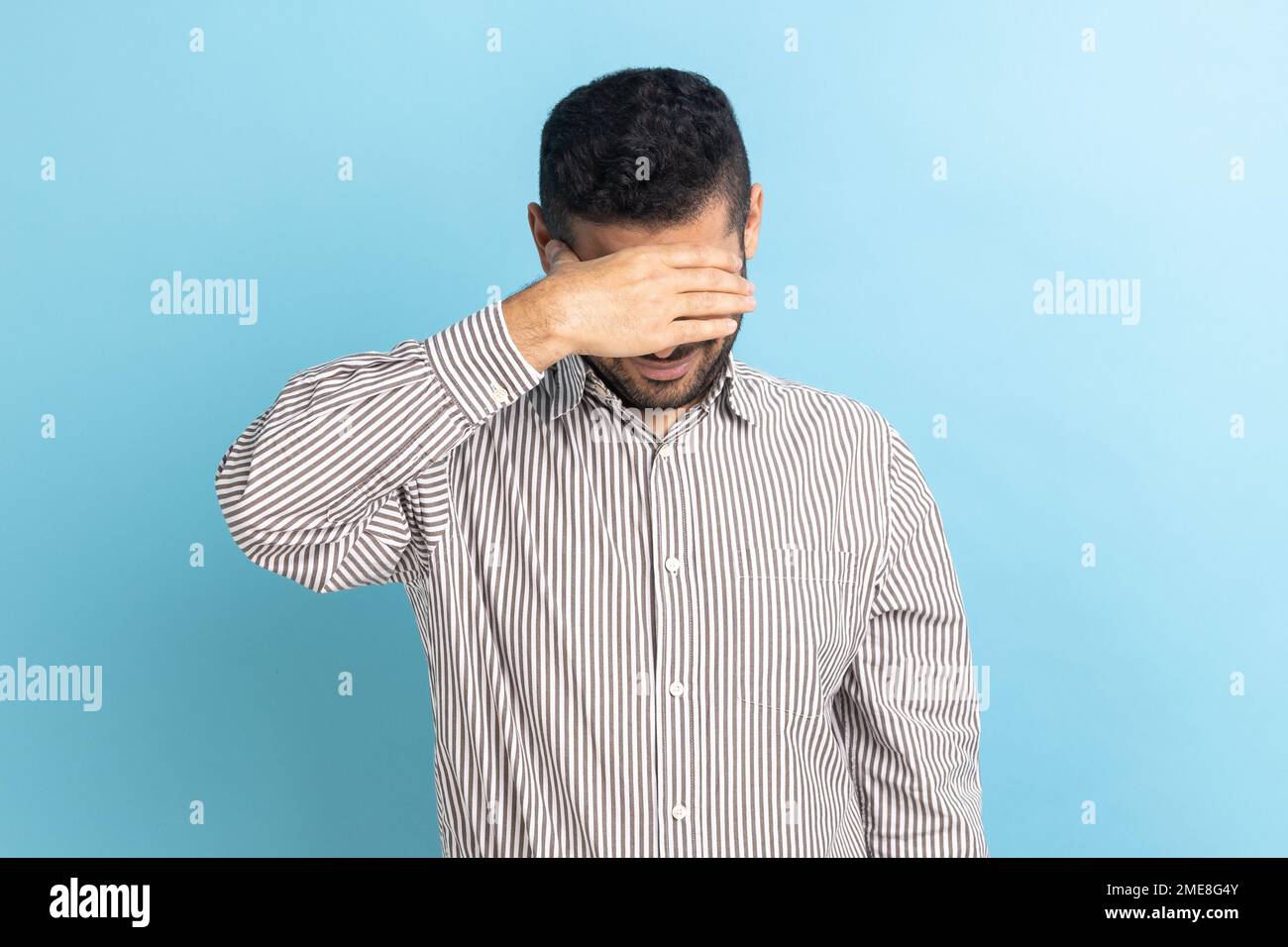 Young adult businessman closing eyes with hand, dont want to see that, ignoring problems, hiding from stressful situations, wearing striped shirt. Indoor studio shot isolated on blue background. Stock Photo
