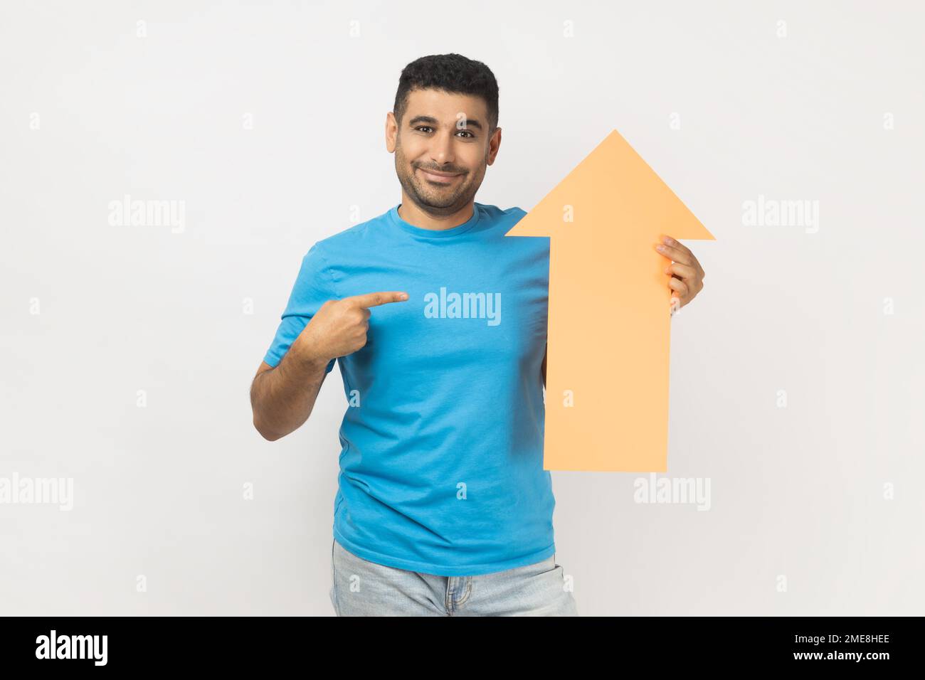 Portrait of cheerful unshaven man wearing blue T- shirt standing pointing at carton arrow indicating up, being glad of career success. Indoor studio shot isolated on gray background. Stock Photo