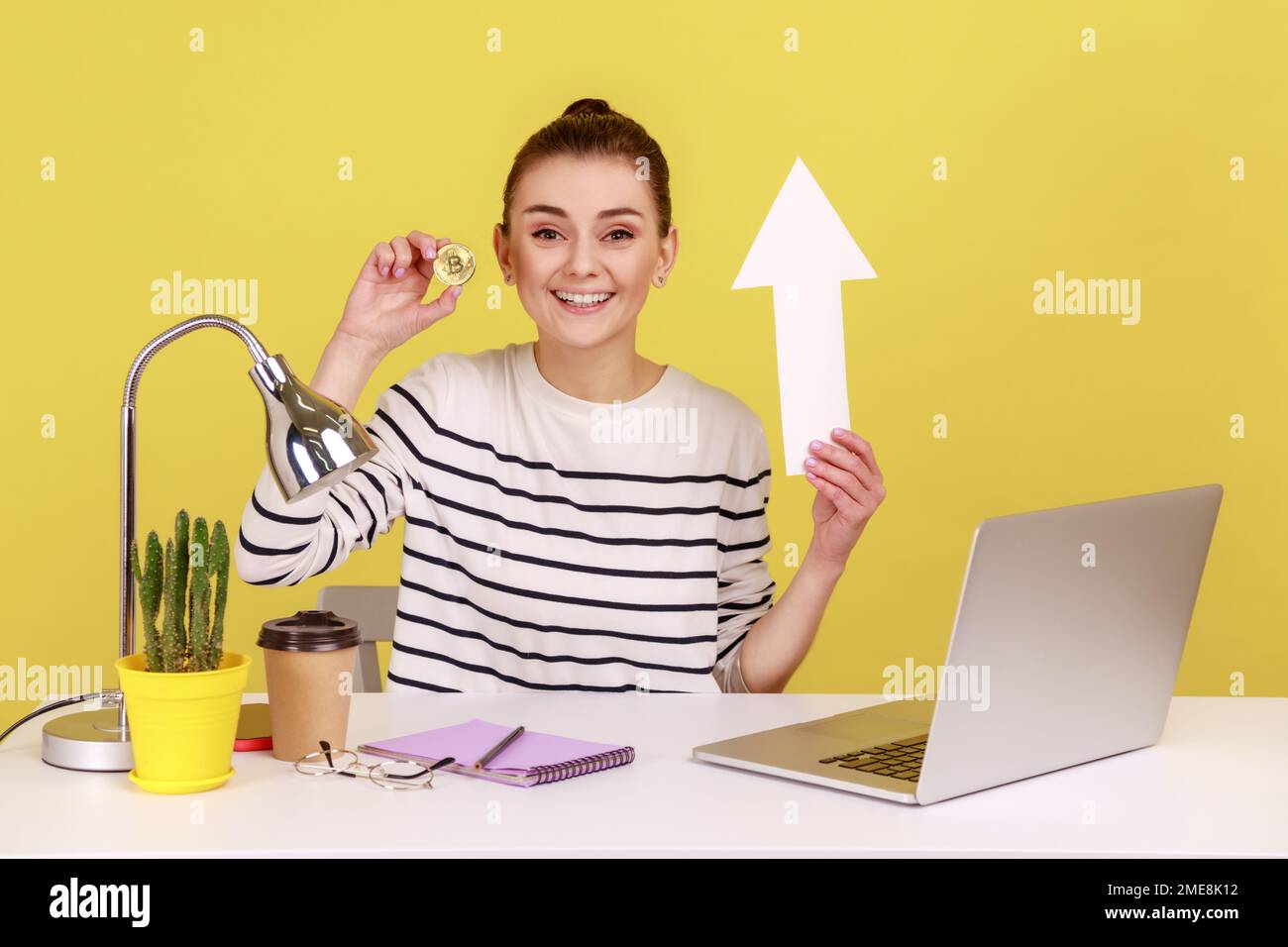 Portrait of happy optimistic woman holding golden bitcoin and white paper arrow, showing growth of crypto currency, sitting on workplace. Indoor studio studio shot isolated on yellow background. Stock Photo