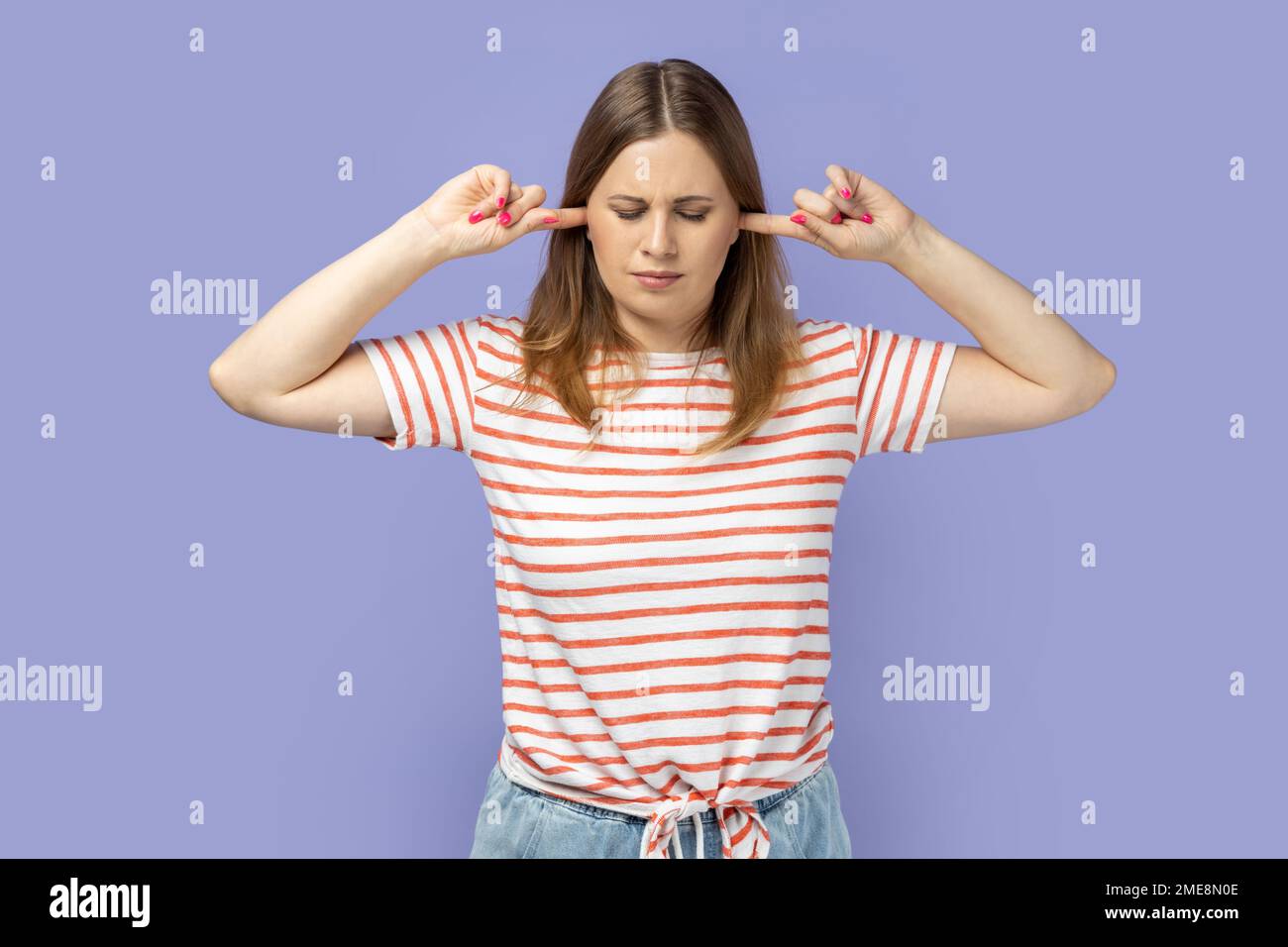 Portrait of depressed attractive blond woman wearing striped T-shirt standing covering ears with fingers, being irritated by noise, closed her eyes. Indoor studio shot isolated on purple background. Stock Photo