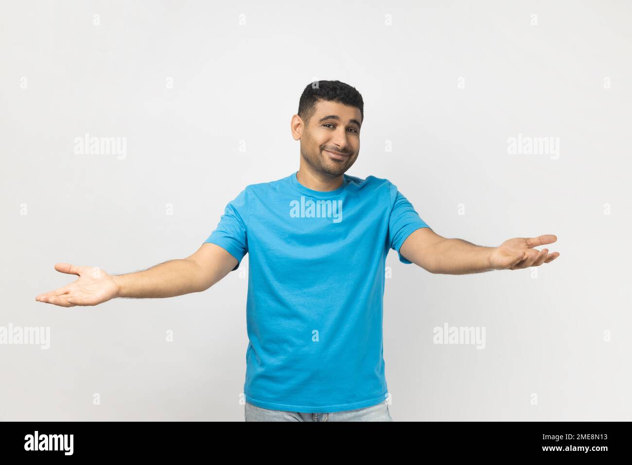 Portrait of unshaven man wearing blue T- shirt standing shows welcome gesture, spreads hands as wants to cuddle best friend, smiles broadly. Indoor studio shot isolated on gray background. Stock Photo