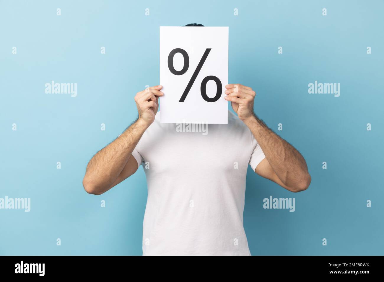 Portrait of anonymous unknown man wearing white T-shirt hiding his face behind paper with percent sign inscription. Indoor studio shot isolated on blue background. Stock Photo