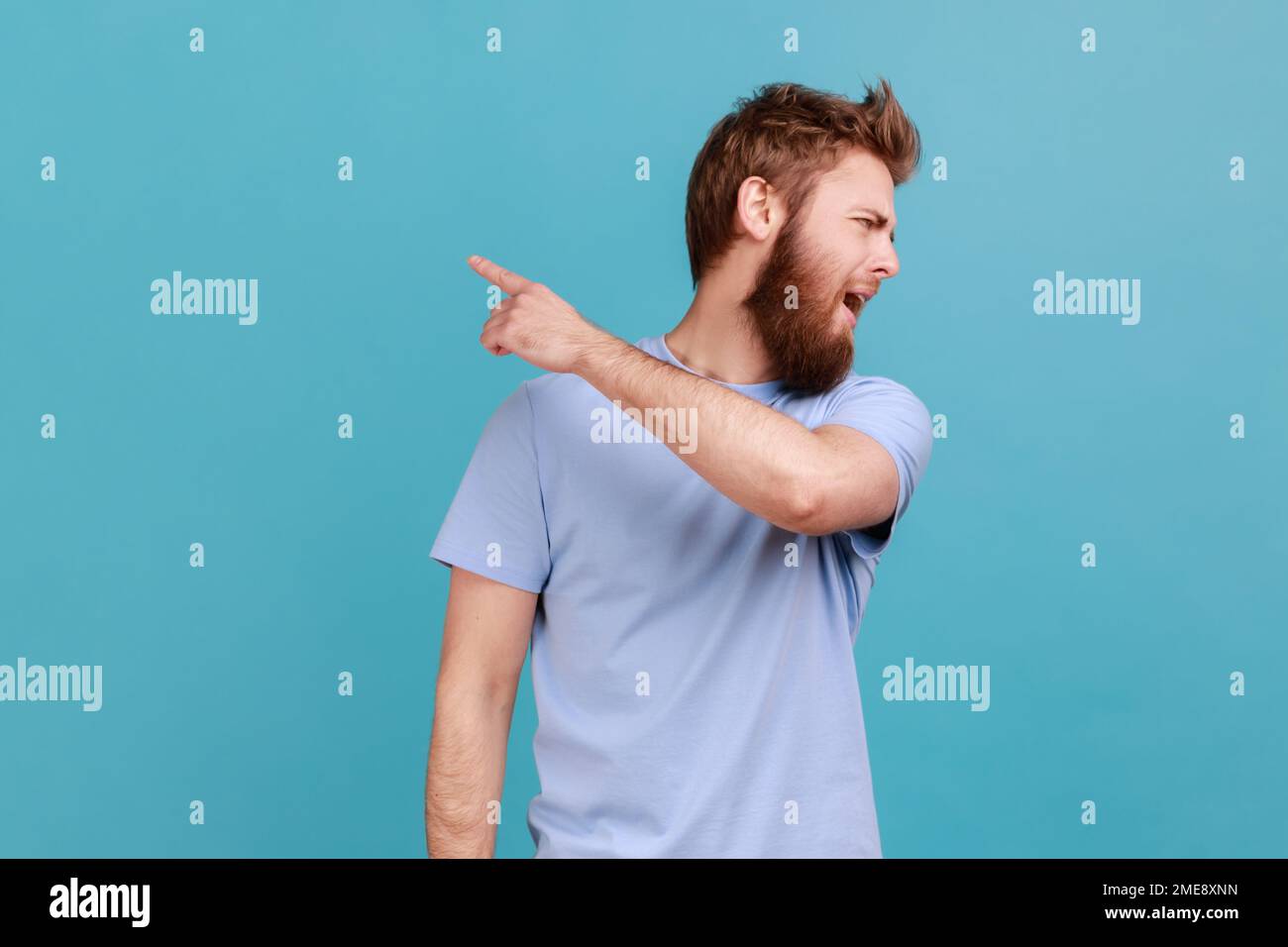 Get out. Portrait of handsome bearded man looking aside and pointing finger another way, asking to leave, dissatisfied expression. Indoor studio shot isolated on blue background. Stock Photo