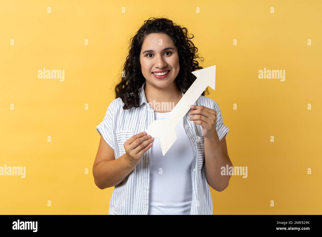 Portrait of smiling satisfied woman with dark wavy hair pointing direction with arrow aside, looking at camera with positive expression. Indoor studio shot isolated on yellow background. Stock Photo