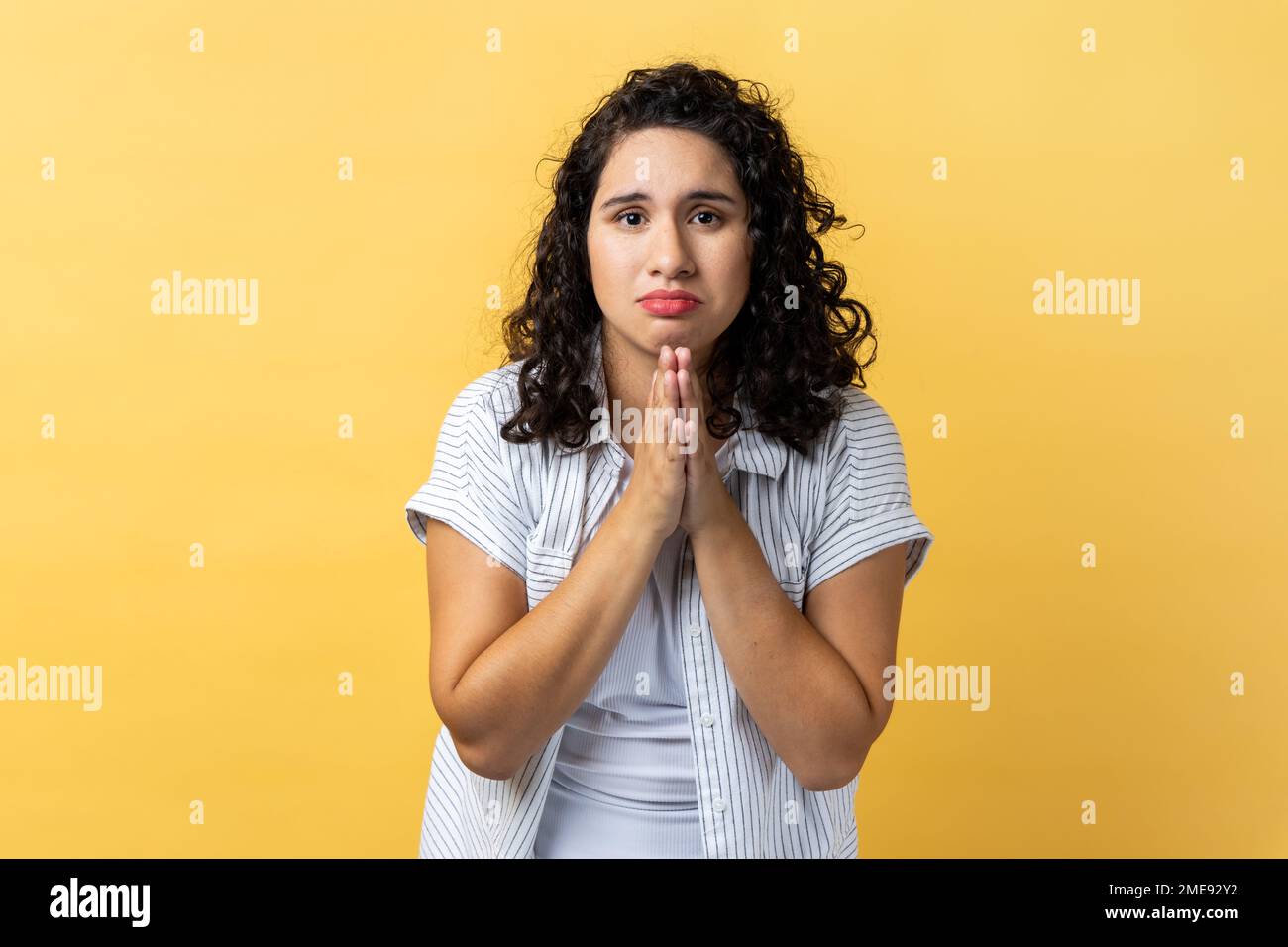 Portrait of woman with dark wavy hair holding hands in prayer, looking with imploring pleading expression, begging help, asking forgiveness. Indoor studio shot isolated on yellow background. Stock Photo