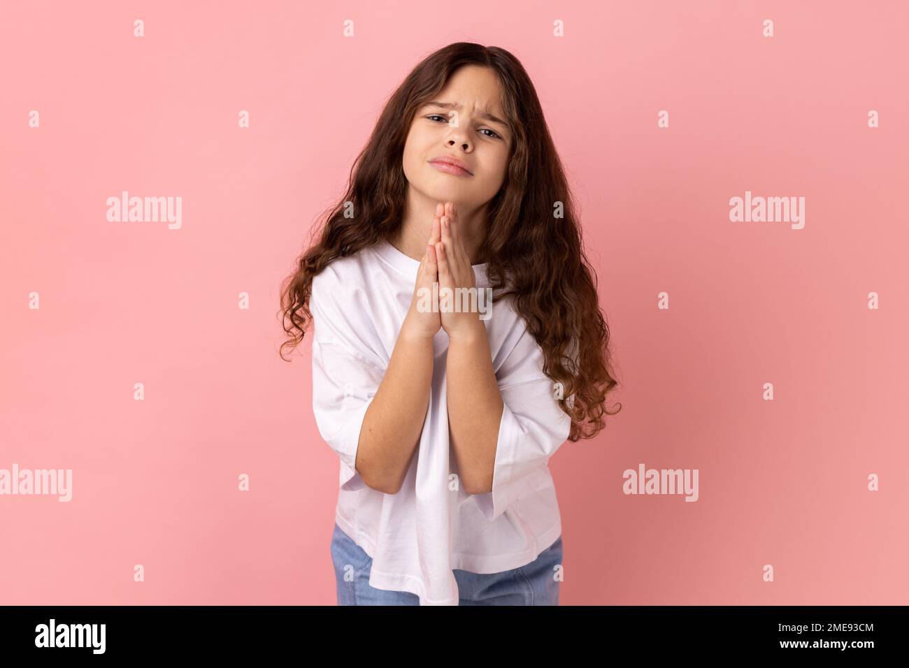 Please help me or forgive. Portrait of little girl wearing white T-shirt standing with palm hands and looking at camera pleased pleading. Indoor studio shot isolated on pink background. Stock Photo