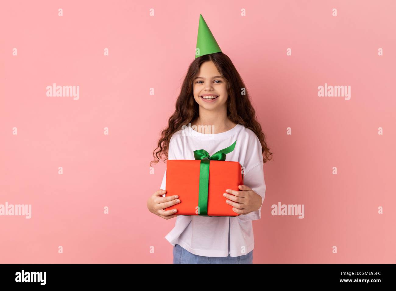 Portrait of little girl wearing white T-shirt and party cone holding red wrapped gift box, looking at camera with satisfied expression. Indoor studio shot isolated on pink background. Stock Photo