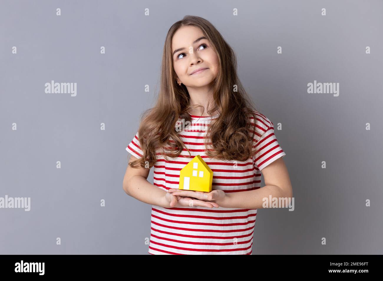 Portrait of little girl wearing striped T-shirt holding in hands little paper house looking away with dreamy expression, new apartment. Indoor studio shot isolated on gray background. Stock Photo