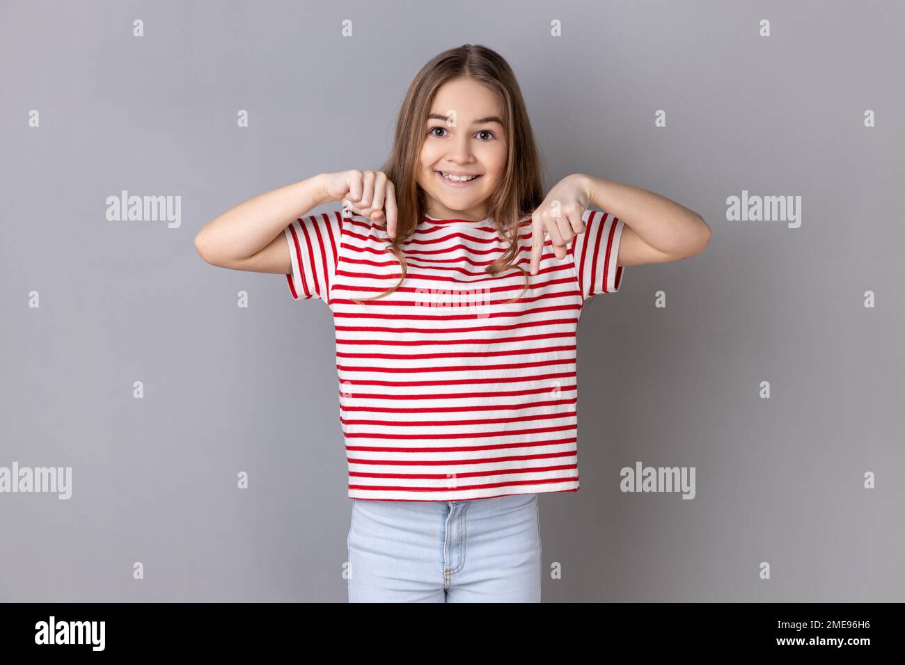 Look, advertise below. Portrait of little girl wearing striped T-shirt pointing down place for commercial idea, looking at camera with toothy smile. Indoor studio shot isolated on gray background. Stock Photo