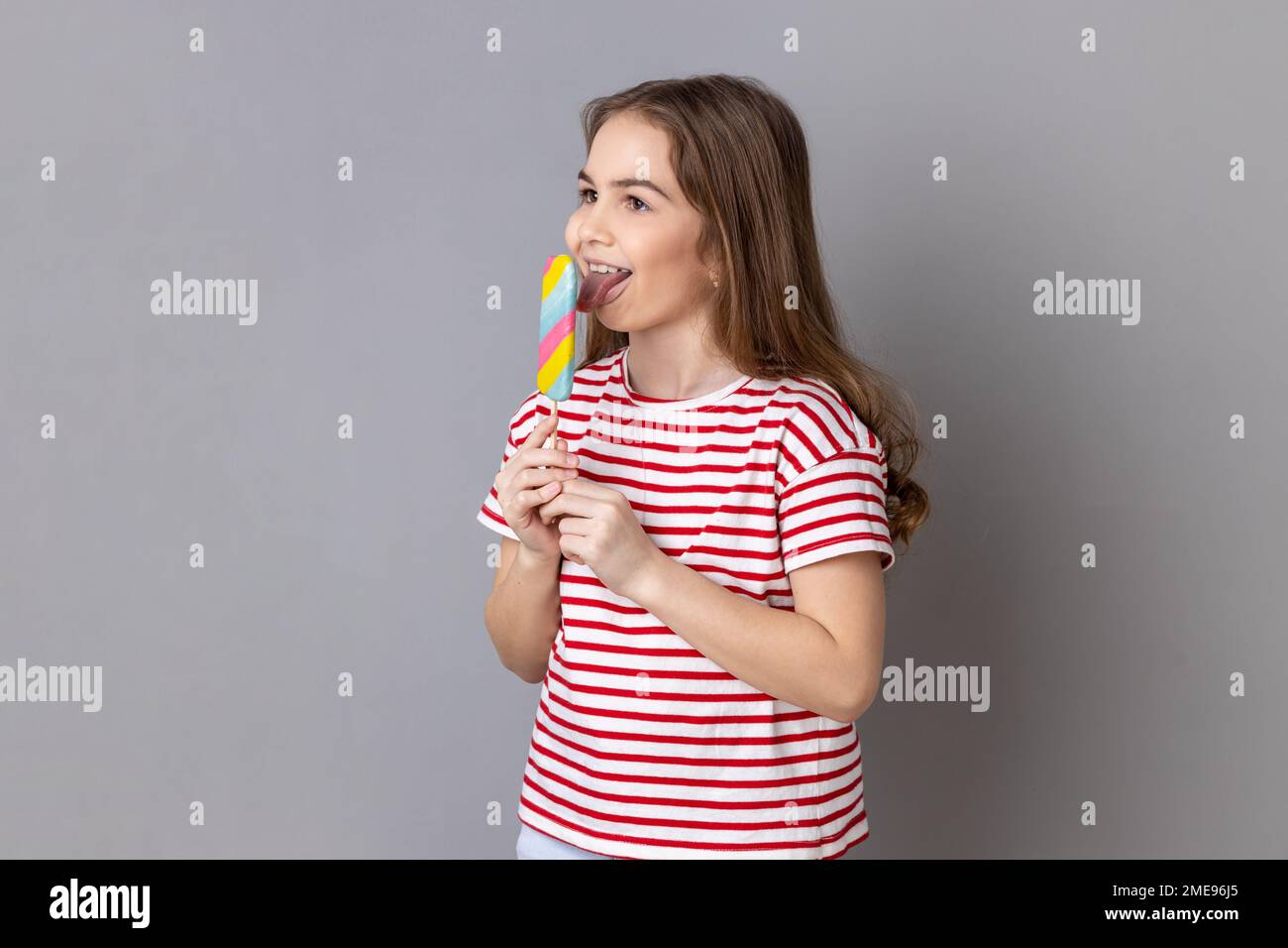 Portrait of little girl wearing striped T-shirt holding colorful candy, licking, looking away with childish satisfied expression. Indoor studio shot isolated on gray background. Stock Photo