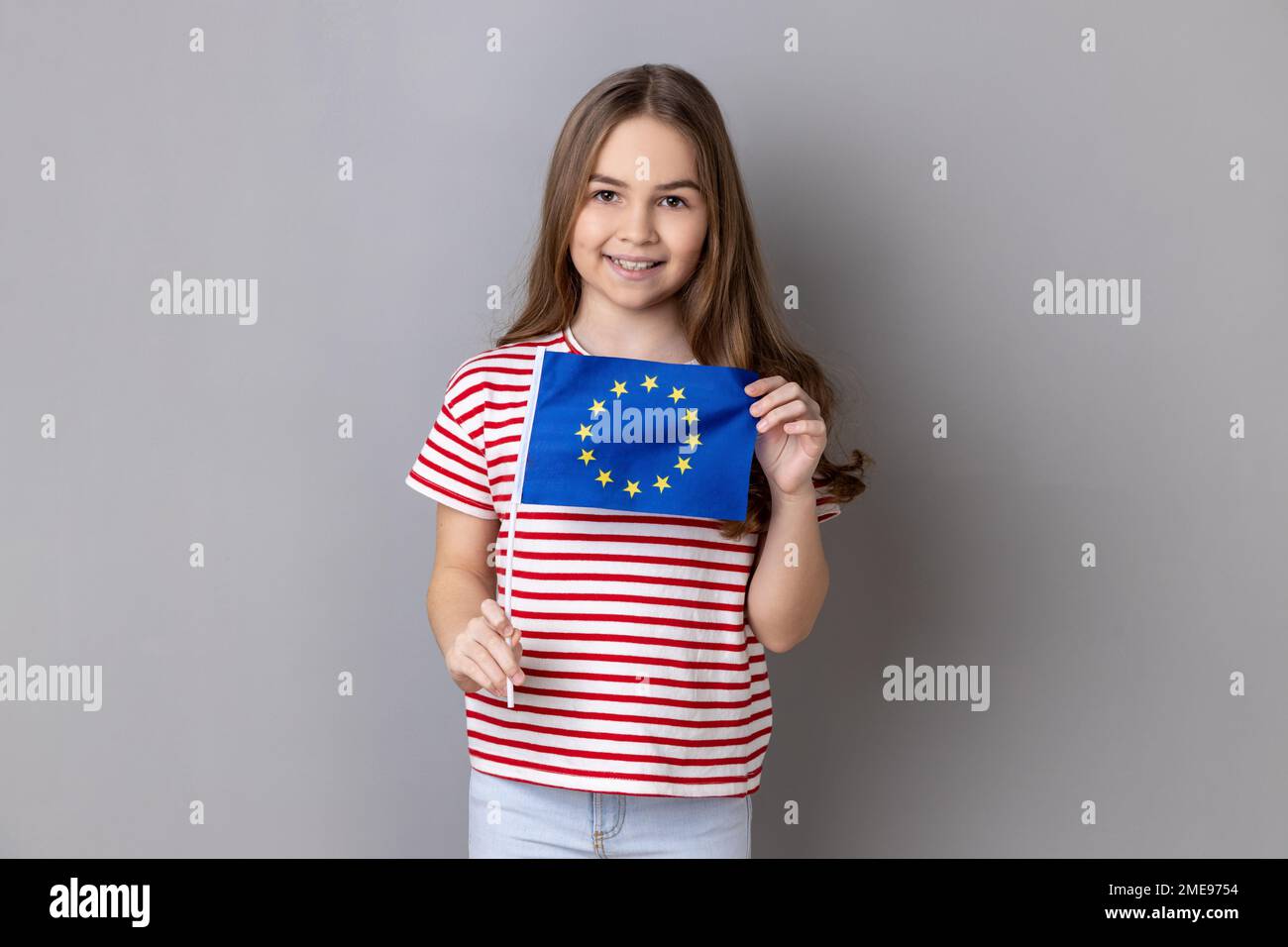 European Union flag. Portrait of smiling adorable cute little girl wearing striped T-shirt holding Europe flag, looking at camera. Indoor studio shot isolated on gray background. Stock Photo