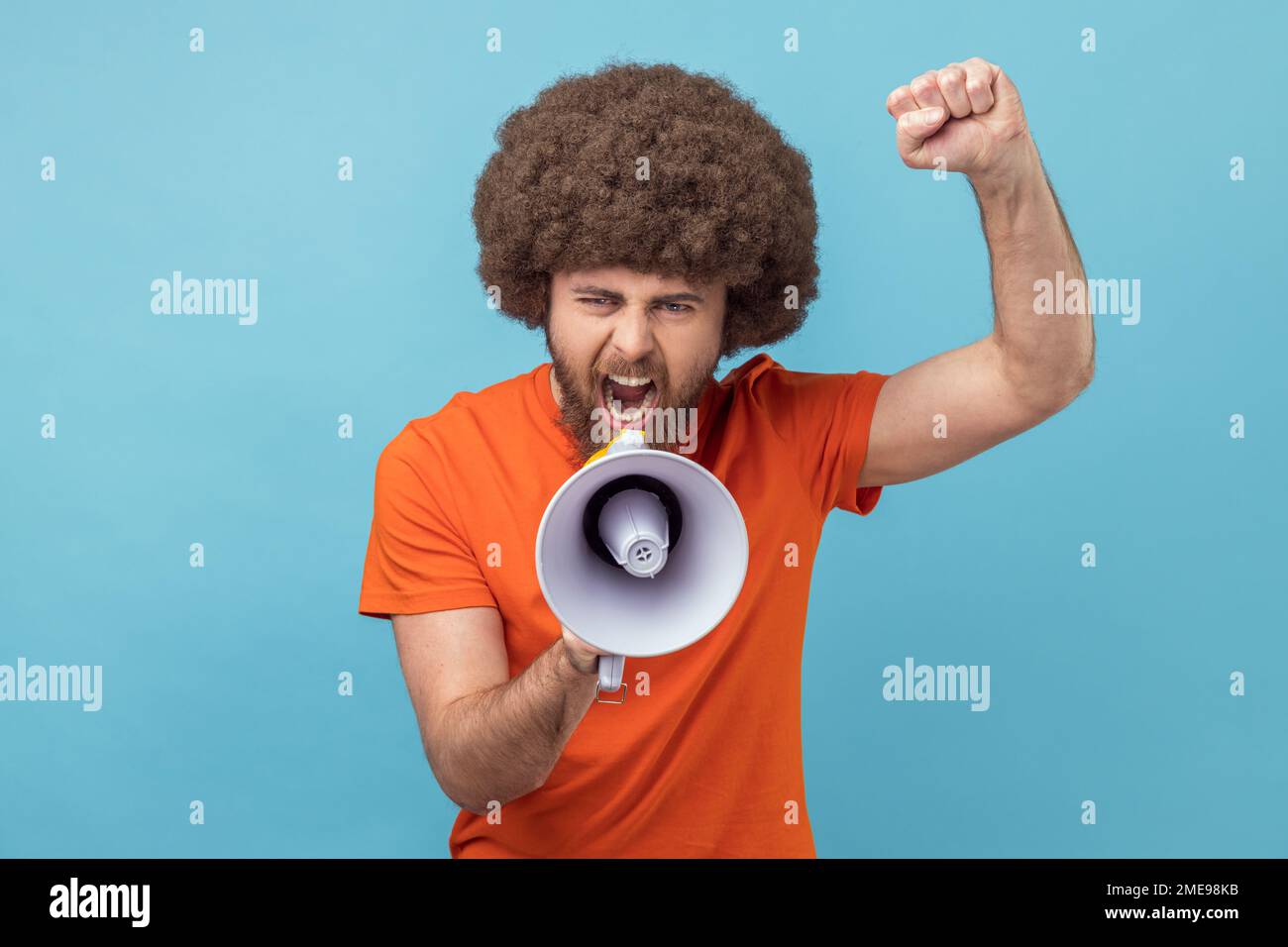 Portrait of serious man with Afro hairstyle wearing orange T-shirt raised hands and holding megaphone, screaming in loud speaker, protesting. Indoor studio shot isolated on blue background. Stock Photo