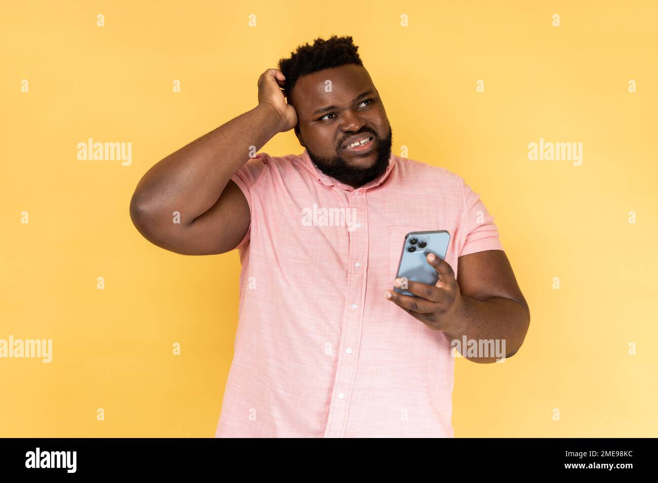 Portrait of pensive thoughtful bearded man wearing pink shirt standing with mobile phone, touching head, having doubt suspicion feeling. Indoor studio shot isolated on yellow background. Stock Photo