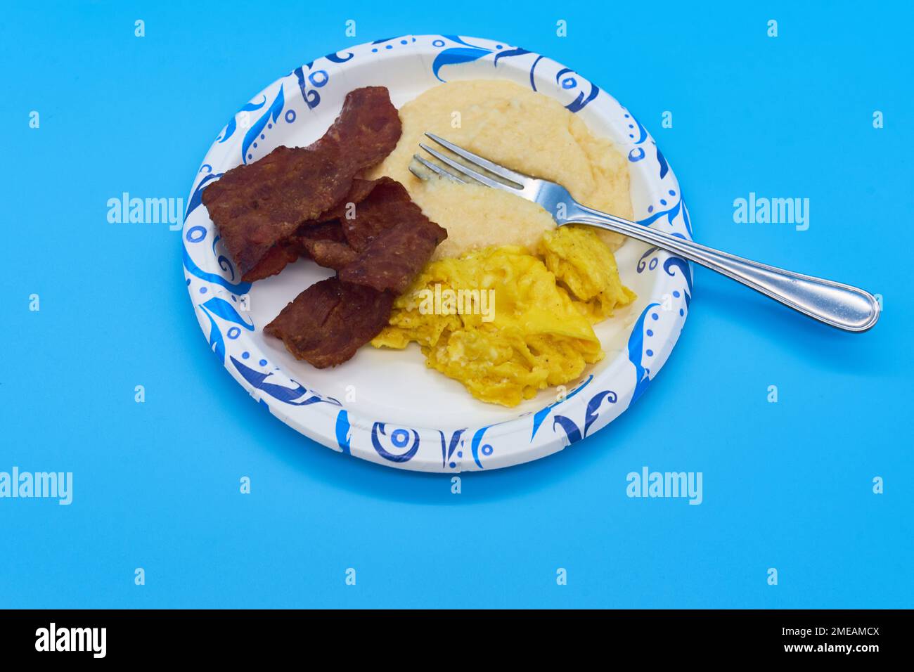 breakfast turkey bacon grits and scrambled eggs on paper plate and light blue background Stock