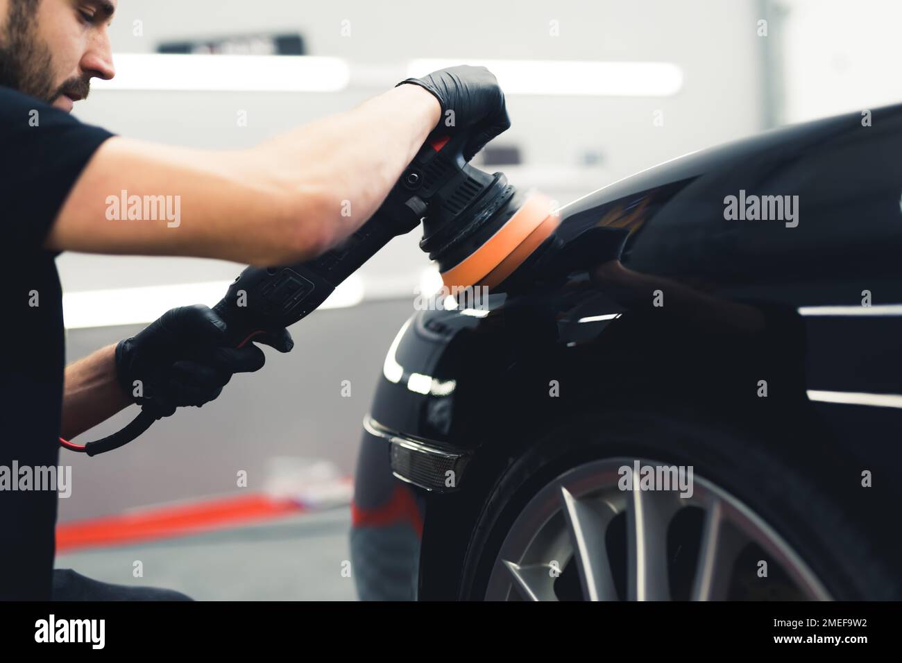 Caucasian man wearing black gloves crouching by the front of a black car polishing it using