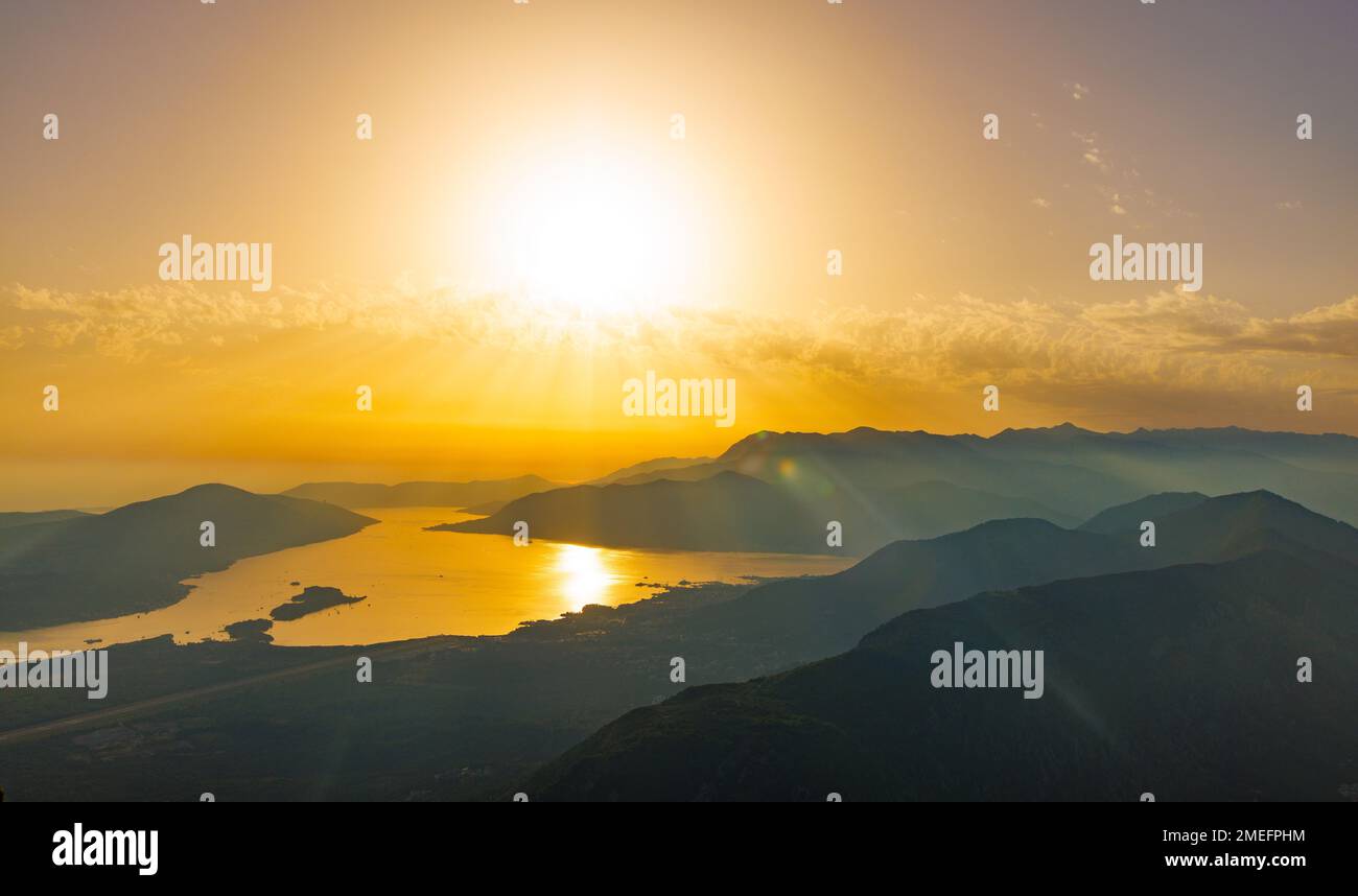 Bright dazzling golden summer sun in the blue cloudy evening sky illuminates all the silhouettes of the peaks of the Balkan Montenegrin mountains and Stock Photo