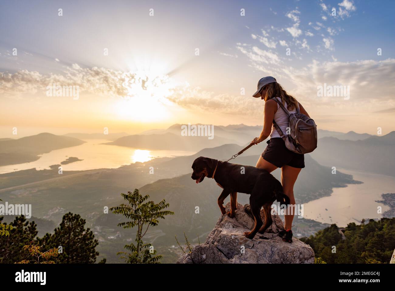 A young happy girl with a backpack stands with her faithful dog Rottweiler on top of a high Montenegrin mountain against the backdrop of coastal citie Stock Photo