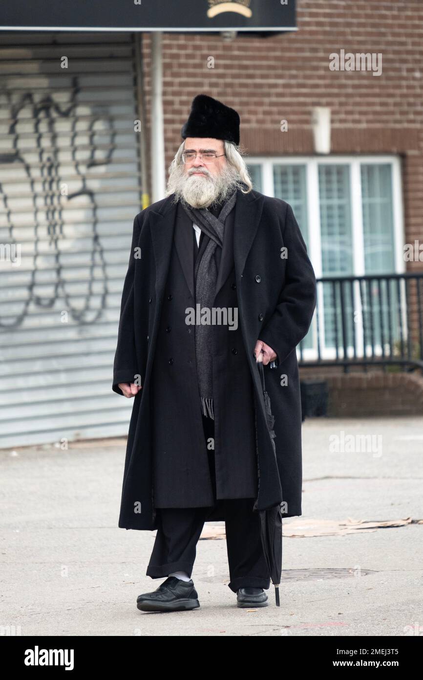 On an overcast day, an older Jewish man  with white gray hair & wearing a karakul hat and carrying an umbrella walks slowly. In Brooklyn, New York Stock Photo