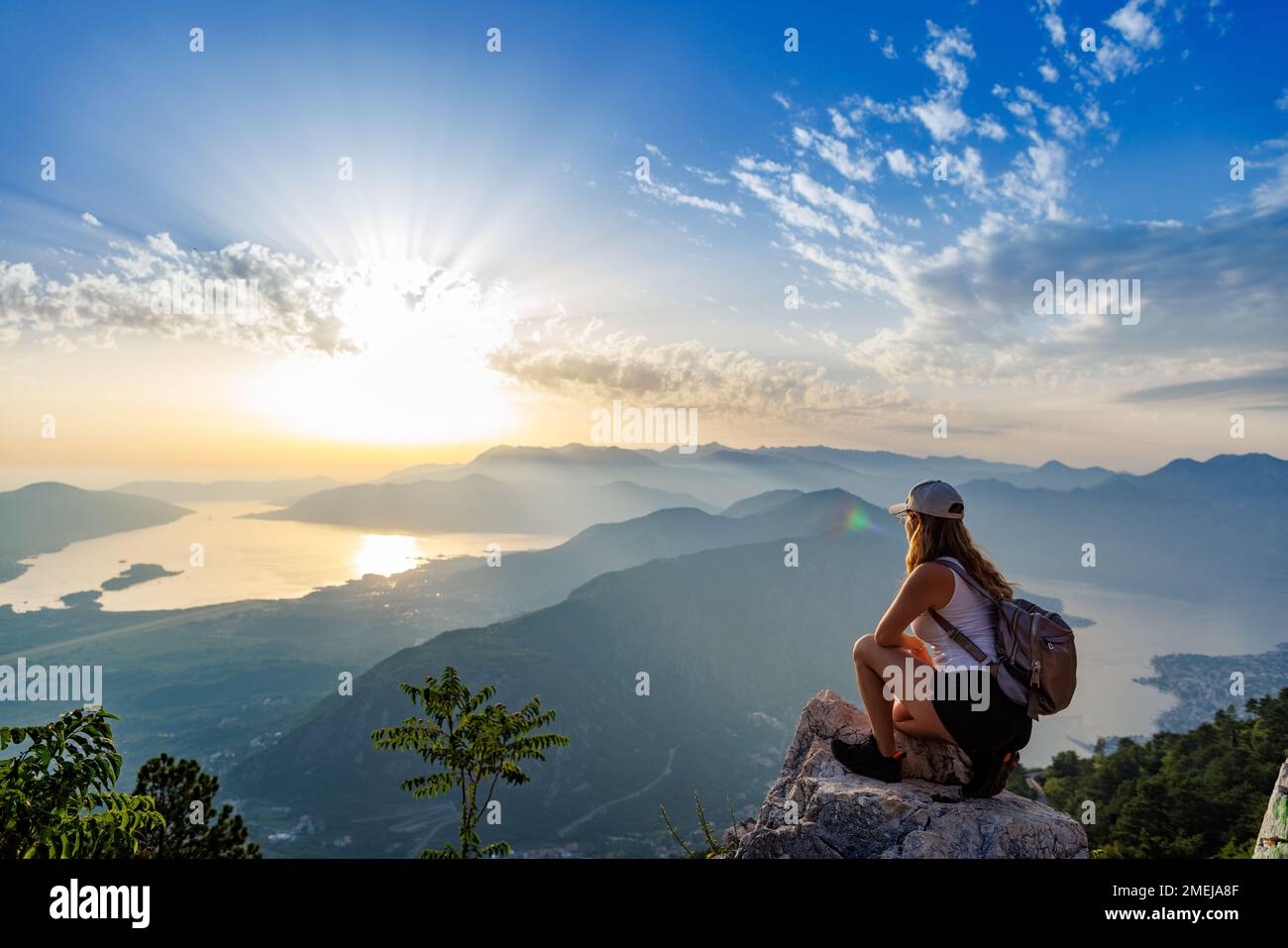 Happy satisfied young girl with a backpack looks at the coastal sea Montenegrin cities, the Adriatic Sea and the bright sunset rays from the top of a Stock Photo