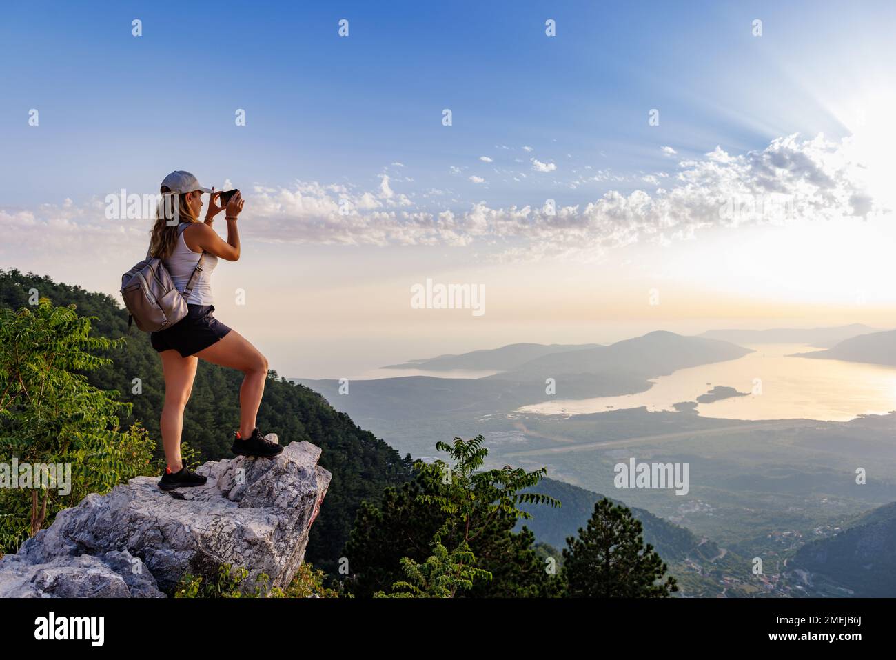 A happy contented young girl with a backpack takes a souvenir photo of the Montenegrin coastal cities, the Adriatic Sea and the bright sunset rays fro Stock Photo